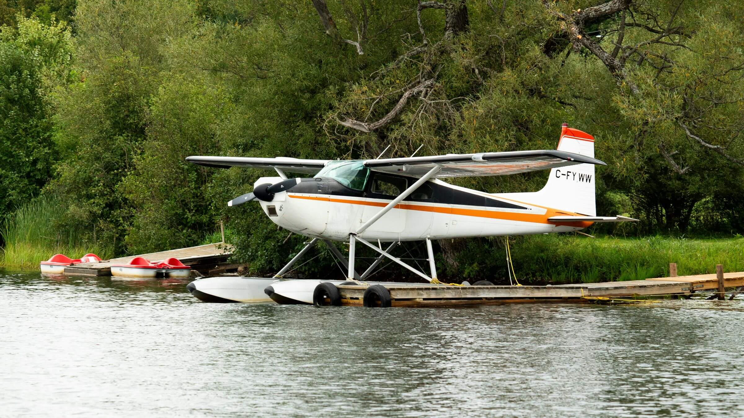 A floatplane is docked by the shore of a lake, surrounded by lush green trees. Two red pedal boats are also moored nearby, contributing to a peaceful lakeside scene.