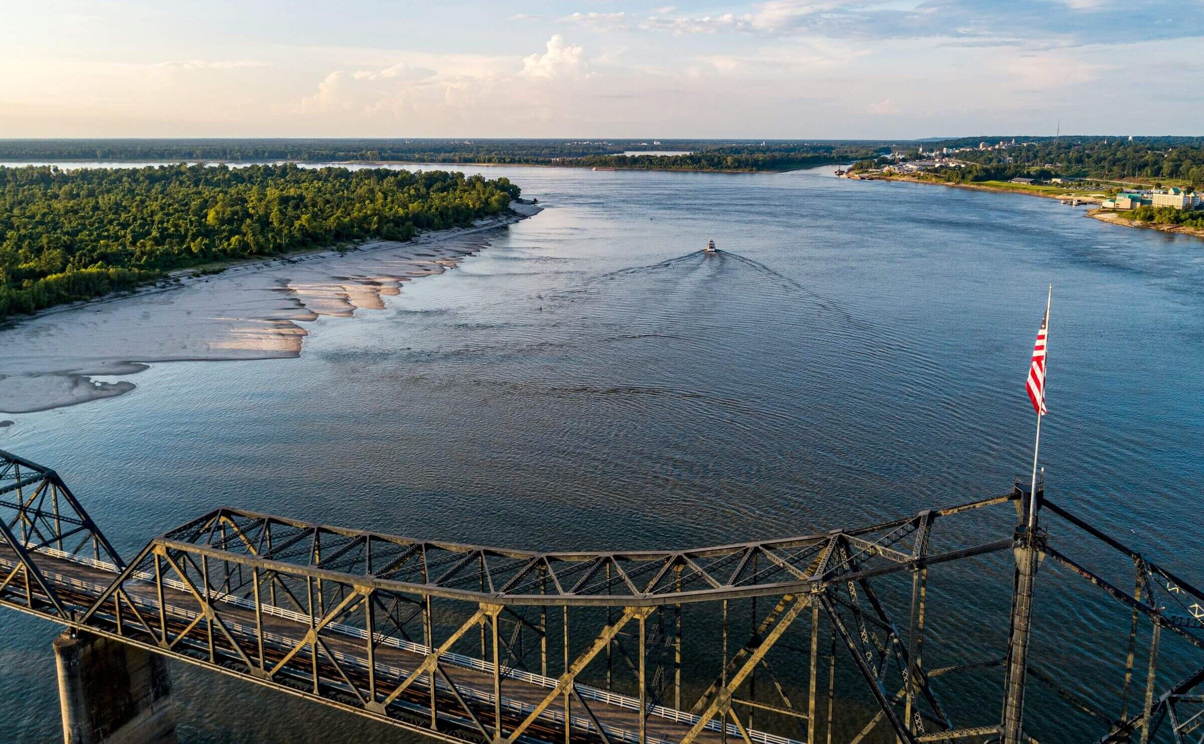 Aerial view of a river with a bridge in the foreground and a boat traveling upstream. Lush green trees line the left bank, and the sky is partly cloudy. An American flag is visible on the bridge.