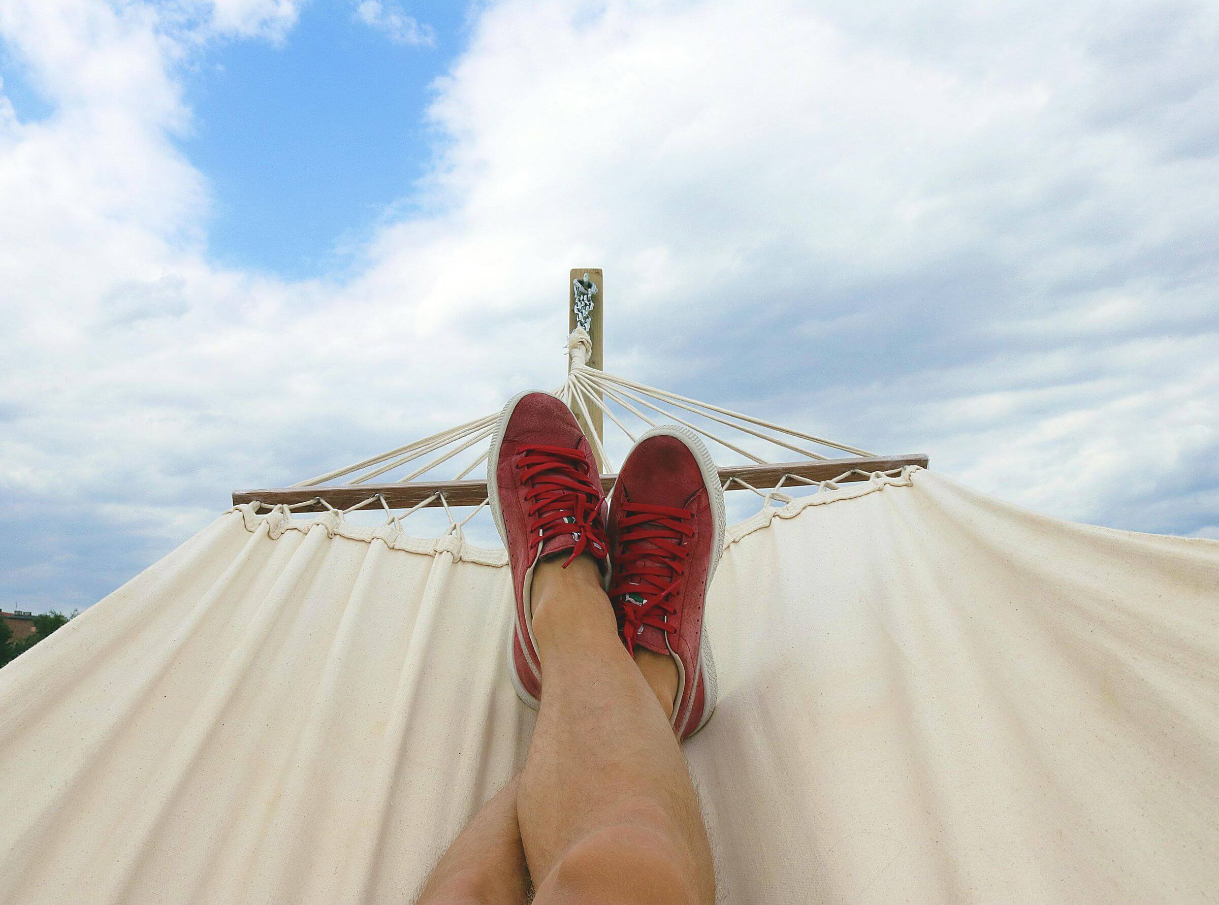 Person relaxing in a hammock under a partly cloudy sky. The view focuses on their crossed legs wearing red sneakers, with the hammock stretching toward the sky.