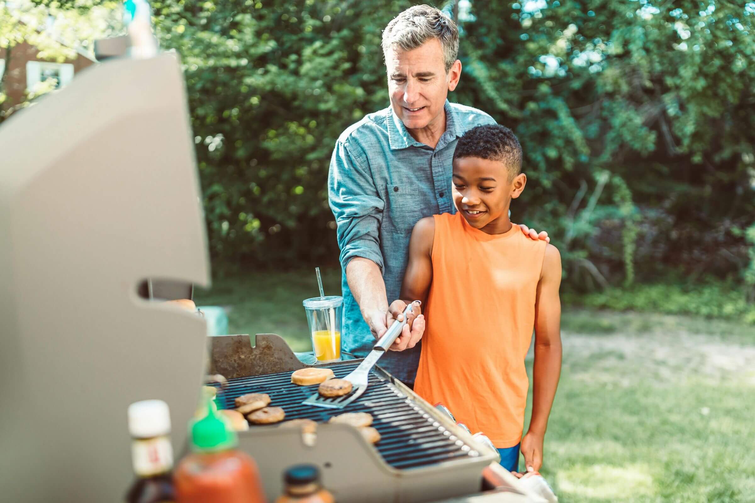 A man and a child are grilling burgers outside on a sunny day. The man guides the child, who is holding a spatula, as they cook on a barbecue. Various condiments are seen in the foreground. Greenery is visible in the background.