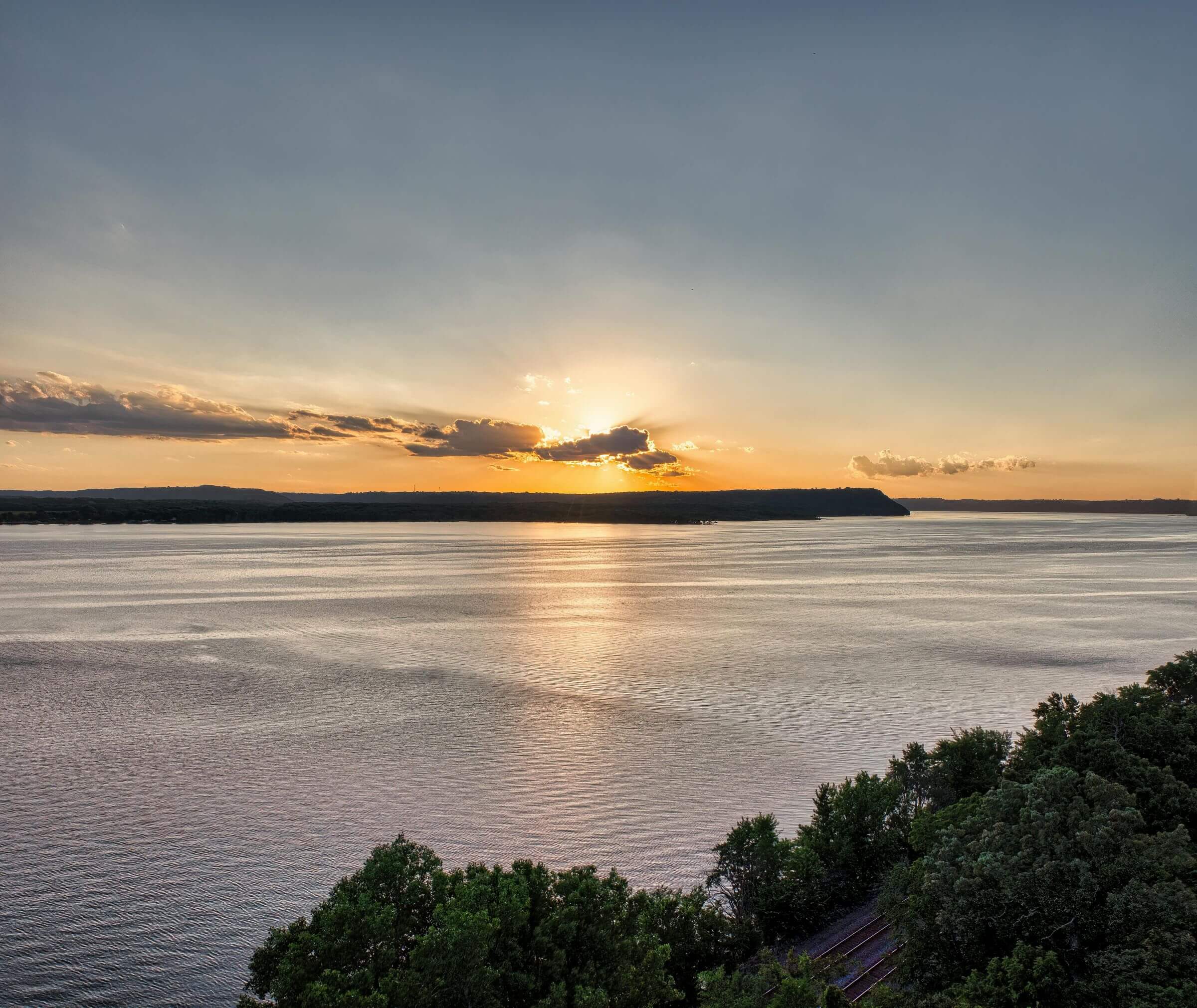 A serene sunset over a vast body of water, with the sun partially hidden by clouds. The sky displays hues of orange and blue as the sun reflects on the water's surface. In the foreground, lush green trees line the edge of the scene.