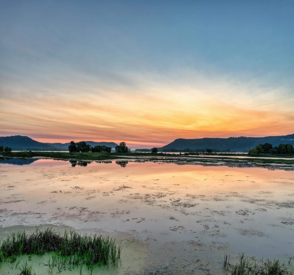 A serene landscape at sunset with a calm lake reflecting the colorful sky. The horizon features silhouettes of distant hills and trees, while the foreground showcases patches of greenery and tranquil water.